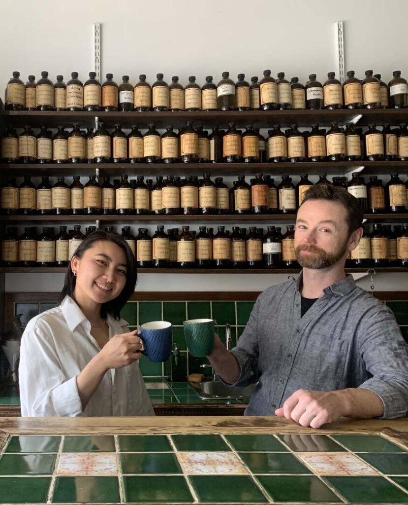 dr. suki hon, ND and dr. chris pickrell holding teacups in front of roncesvalles apothecary front desk with tincture bottles in background