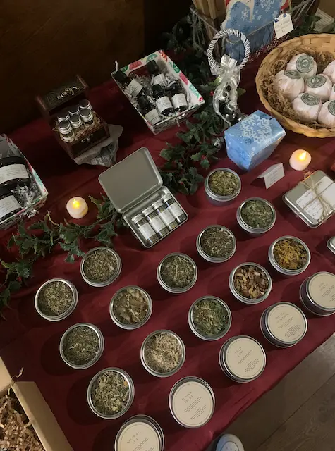various stocking stuffer gifts with herbal tea sample tins, apothecary kits, essential oils, bath bombs laid out on maroon table cloth